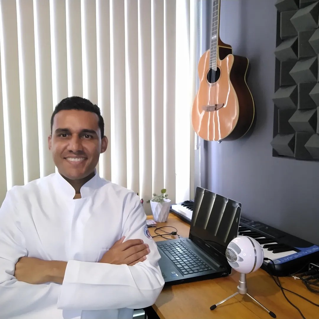 Foto do fonoaudiólogo Gabriel do Nascimento, de jaleco branco, sentado em seu consultório